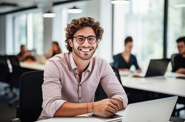 A smiling young man in an office setting, surrounded by colleagues working at their desks, representing a positive work environment