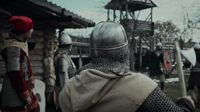 Medium rear shot of unrecognizable male medieval warrior in chainmail and helmet, holding battle axe on shoulder, watching troop mates training with swords outdoors in yard of military garrison