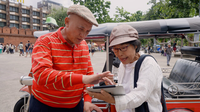 Elderly tourists interacting with local tuk-tuk driver in Chiang Mai, Thailand. Man in red shirt and woman in hat looking at tablet. Scene of tourists receiving guidance and enjoying cultural exchange
