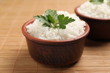 Bowls of delicious boiled rice with parsley on bamboo mat, closeup