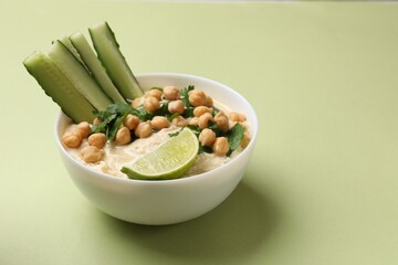 Tasty hummus with chickpeas, cucumber, lime and parsley in bowl on light green table. Space for text