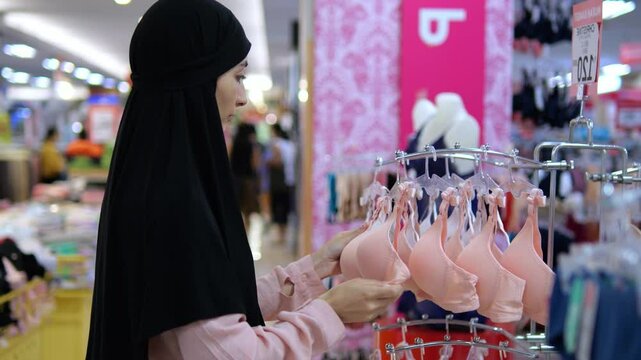 Muslim woman in hijab chooses underwear in clothing store. Woman carefully considers different models underwear, taking into account preferences. Choice underwear combination tradition and modernity.