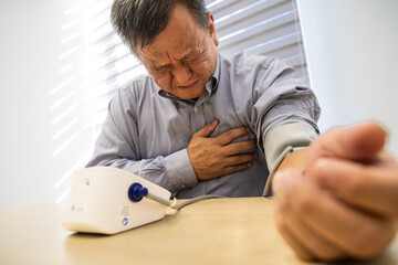 Stressful Asian man in 50s with painful chest checking blood pressure with instrument in the work...