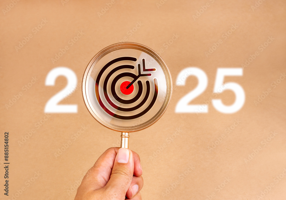 Wall mural Happy new year 2025 with business trends concept banner. Target icon inside golden magnifying glass holding by hand with white defocused 2025 year number on brown background. Searching goal concept.