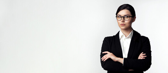 A woman in a black suit and glasses stands with her arms crossed in front of a white background.