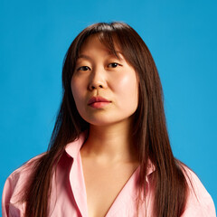 Portrait of confident Asian woman with brunette hair in pink shirt posing with serious and calm expression against blue studio background. Concept of human emotions, youth, lifestyle