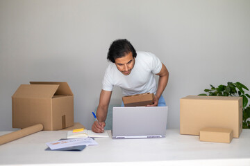 Man working on his laptop and taking notes, holding a box, managing a small business.