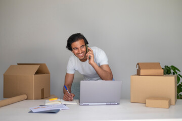 Smiling man on the phone, working on his laptop and writing notes, managing a small business.
