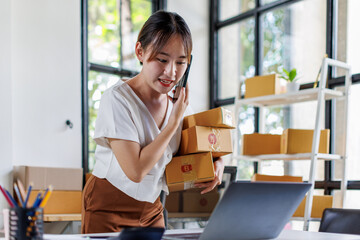 Young asia seller woman happy work on laptop busy cellphone call chat to customer at home office desk use digital tablet computer in SME online store social e-commerce sale page service web support.
