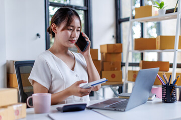 Obraz premium Young asia seller woman happy work on laptop busy cellphone call chat to customer at home office desk use digital tablet computer in SME online store social e-commerce sale page service web support. 