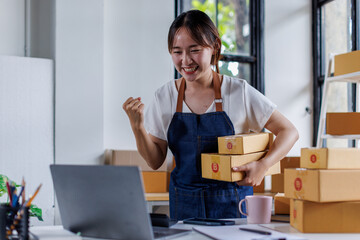 Happy young asian woman startup small business freelance holding parcel box and computer laptop and sitting on chair, Online marketing packing SME box delivery concept