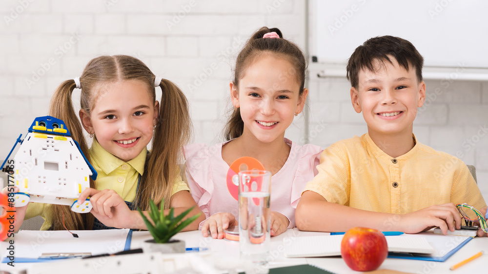 Poster Three children, a girl holding a robot and two others, are sitting at a table in a classroom setting, smiling and working on a STEM project.