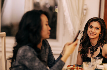 Two attractive multiethnic female friends in evening dresses are sitting at a festive table. A charming Asian woman writes messages on her phone.