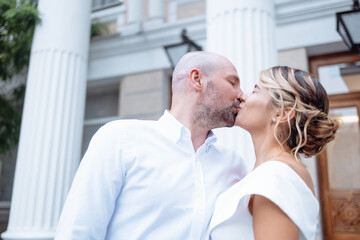 Attractive groom kisses charming bride against background of building with large columns. Young man in white shirt embraces beautiful woman in elegant dress.