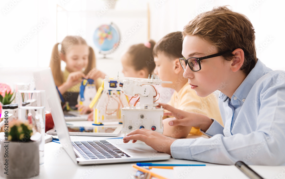 Poster a teenage boy wearing glasses sits at a desk in a classroom, using a laptop computer. he is surround