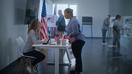 African American woman comes to registration table at polling station. Female polling worker talks to woman and uses digital tablet. Multiethnic US citizens vote in voting booths. Election Day in USA.