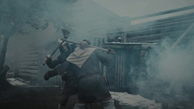 Medium full slowmo shot of two male warriors clad in chainmail, armour and helmets having violent fight with battle axes in courtyard of burning wooden castle, in plumes of smoke, during feudal war