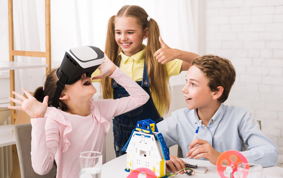 Three children are working on a robotics project. One girl is wearing a virtual reality headset and has a look of excitement on her face. The other two children are watching her,