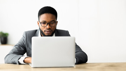 A man in a suit and glasses is working on a laptop at his desk in an office.