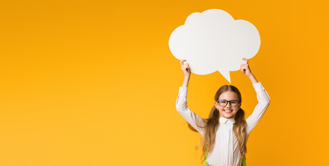 Smiling Elementary Student Girl Holding Speech Bubble Overhead On Yellow Studio Background. Thinking Of School. Mockup, Free Space