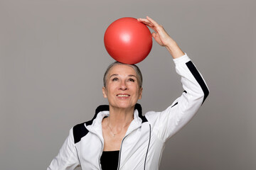 Happy active senior woman balancing red ball on head against gray background
