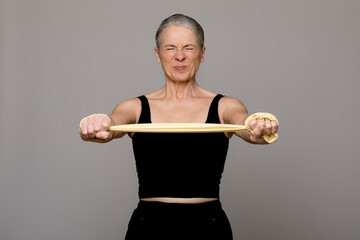 Senior woman exercising with resistance band grimacing against gray background