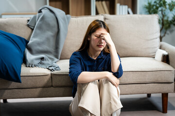 A young Asian woman sits on a sofa in her living room, feeling sick with mental health diseases. She struggles with depression, anxiety, and other conditions, seeking support and solace at home.