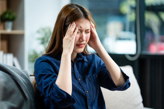 A young Asian woman sits on a sofa in her living room, feeling sick with a headache. She experiences throbbing pain and nausea, seeking comfort and relief at home.