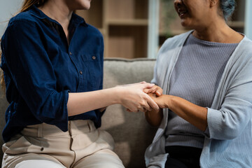 A mature mom and a young Asian woman, mother and daughter, sit together on a sofa in their living room, celebrating Mother's Day with love and bonding at home.