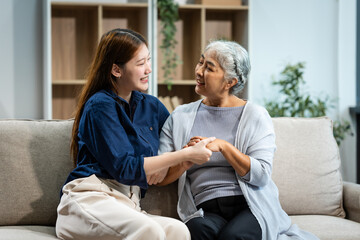 Obraz premium A mature mom and a young Asian woman, mother and daughter, sit together on a sofa in their living room, celebrating Mother's Day with love and bonding at home.