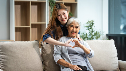A mature mom and a young Asian woman, mother and daughter, sit together on a sofa in their living room, celebrating Mother's Day with love and bonding at home.