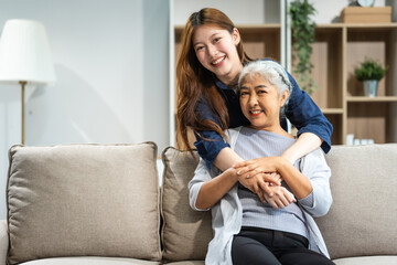 A mature mom and a young Asian woman, mother and daughter, sit together on a sofa in their living room, celebrating Mother's Day with love and bonding at home.