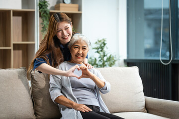 A mature mom and a young Asian woman, mother and daughter, sit together on a sofa in their living room, celebrating Mother's Day with love and bonding at home.