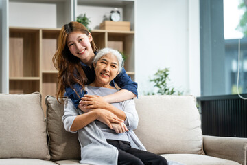 Obraz premium A mature mom and a young Asian woman, mother and daughter, sit together on a sofa in their living room, celebrating Mother's Day with love and bonding at home.