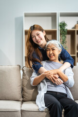 A mature mom and a young Asian woman, mother and daughter, sit together on a sofa in their living room, celebrating Mother's Day with love and bonding at home.