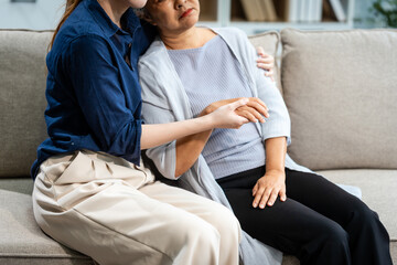 A mature mom and a young Asian woman, mother and daughter, sit together on a sofa in their living room, offering tired consolation and encouragement to each other at home.