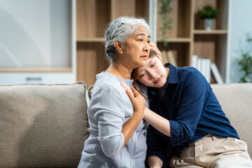 A mature mom and a young Asian woman, mother and daughter, sit together on a sofa in their living room, offering tired consolation and encouragement to each other at home.