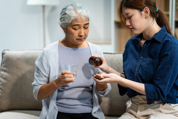 A young Asian woman and her grey-haired mature mom, mother and daughter, sit together on a sofa in their living room. The daughter gives pills and a glass of water to her sick mom.