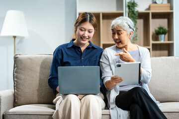 In their living room, a young Asian woman and her grey-haired mature mom, mother and daughter, sit together on a sofa. daughter works online with laptop while mom lovingly cares and supports her.