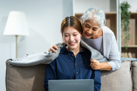 In their living room, a young Asian woman and her grey-haired mature mom, mother and daughter, sit together on a sofa. daughter works online with laptop while mom lovingly cares and supports her.