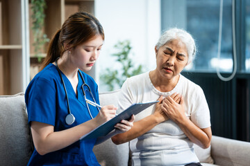 Young Asian nurse attends to older grey-haired mature woman sitting on sofa, conducting health...