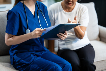Young Asian nurse attends to older grey-haired mature woman sitting on sofa, conducting health...