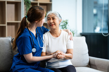 Obraz premium A young Asian nurse, only woman present, holds hands with an older grey-haired mature woman sitting on a sofa, offering encouragement while taking her medical history and conducting health checks.