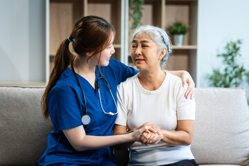 A young Asian nurse, only woman present, holds hands with an older grey-haired mature woman sitting...