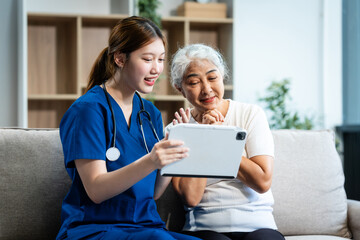 A young Asian nurse cares for an older grey-haired woman on a sofa, assessing health and discussing osteoporosis, arthritis, Alzheimer's, and dementia, using a tablet for information.