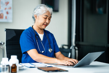 A grey-haired Asian mature woman nurse, doctor, disease specialist, and surgeon works at a desk, utilizing her expertise as an experienced and senior healthcare professional.