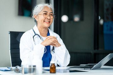 A grey-haired Asian mature woman nurse, doctor, disease specialist, and surgeon works at a desk, utilizing her expertise as an experienced and senior healthcare professional.