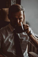 A young man is going to work in the office in the morning in a white shirt and tie having breakfast drinking coffee from a large mug while sitting in an office beige armchair.  Office worker business