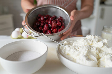 Making a cheesecake with cherries. Woman with apron in the kitchen and ingredients for making a cake.