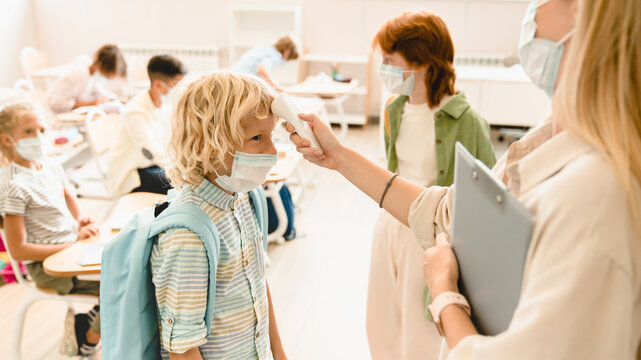Teacher measuring temperature on her student`s forehead classmates schoolchildren before lesson wearing protective medicine face masks against coronavirus Covid19 due to lockdown restrictions. - Powered by Adobe